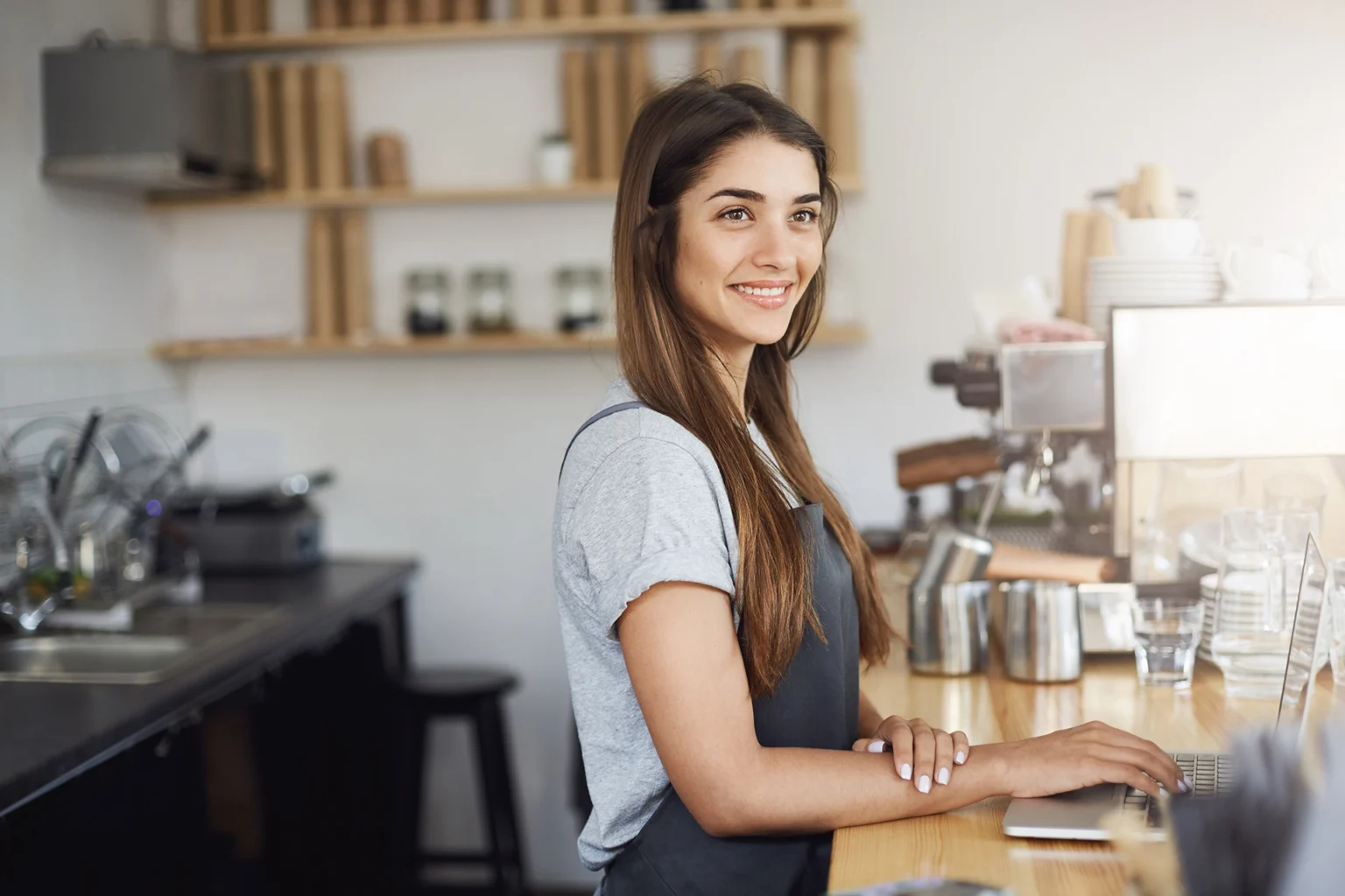 Barista con cabello largo y liso, vestida con camiseta gris y delantal oscuro, de pie detrás de la barra de una cafetería moderna. A su alrededor se observan una máquina de café, vasos transparentes, utensilios de metal, estanterías de madera con frascos y platos apilados al fondo. La iluminación es suave y el ambiente transmite orden y calidez.