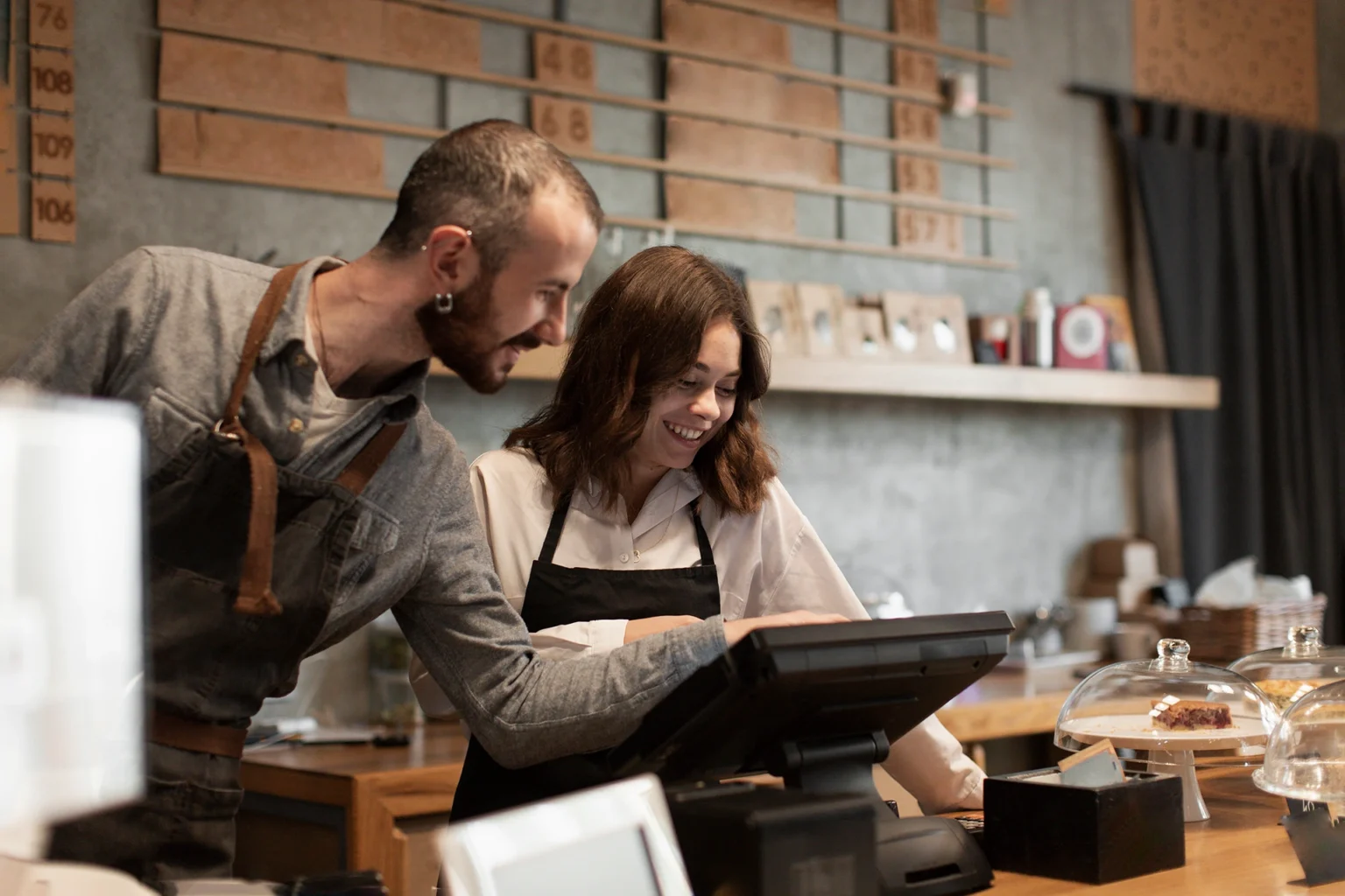 Dos personas con delantales trabajan juntas en la barra de una cafetería, revisando la pantalla de una caja registradora. Alrededor hay vitrinas con pasteles, una estantería con objetos decorativos y un ambiente acogedor con paredes de tono gris.