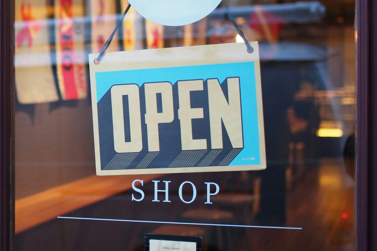 Cartel colgado en la puerta de una tienda con la palabra “OPEN” en letras grandes sobre fondo azul y beige, acompañado del texto “SHOP” en el cristal.