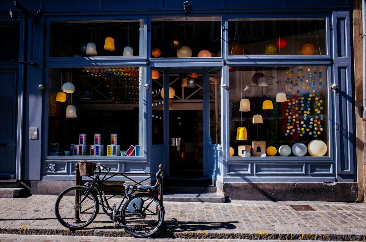 Fachada de una tienda con grandes ventanales que muestran lámparas colgantes y decoraciones de colores en el interior. Una bicicleta negra con cesta está aparcada en la acera frente al escaparate.
